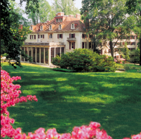 Yellow mansion in the background of a green lawn and pink flowers in the foreground.