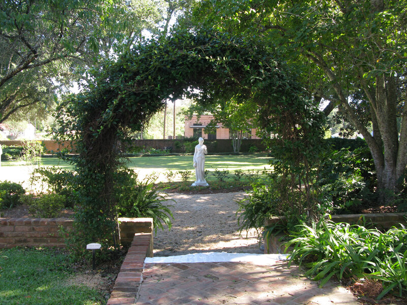 Natural archway over a brick path leading to a white statue of a woman