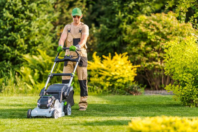 Landscaper in work overalls mowing a bright green lawn in a well-kept garden surrounded by shrubs and sunlit trees.