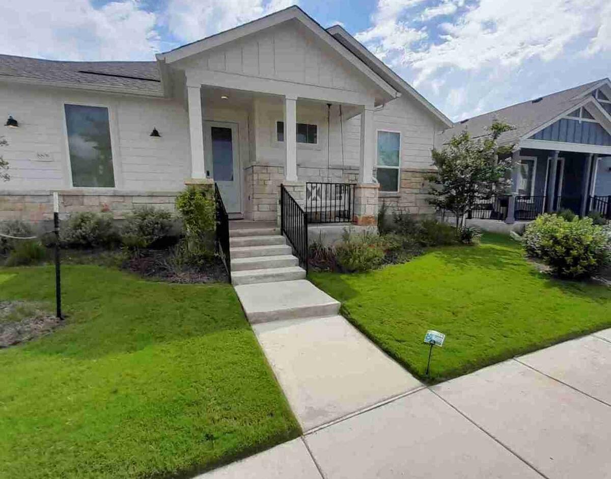 A well-maintained front lawn of a modern suburban home in Austin, Texas, featuring neatly trimmed green grass, landscaped bushes, and a central concrete walkway leading up to a porch with black metal railings.