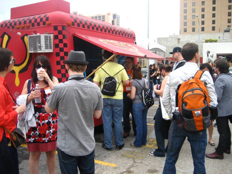 Crowd lines up at red checkered food truck on city street, festivalgoers chat, one woman sips drink nearby.