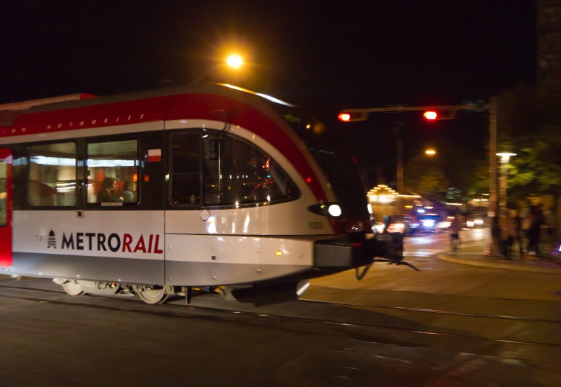 Austin MetroRail train passes through nighttime intersection, headlights and streetlights glowing, cars wait as modern red and silver tram rolls.