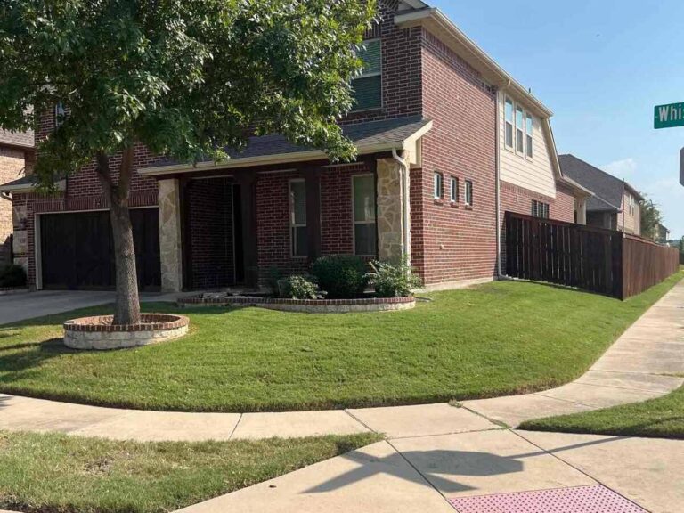 Corner view of a well-maintained red brick house with a manicured lawn, tree in a circular stone planter, and a dark wooden fence along the side. The house is located at the intersection of a residential street with visible sidewalk and street sign