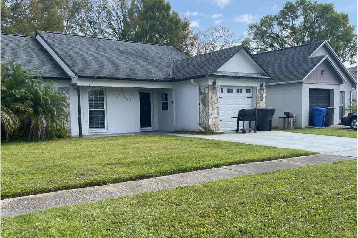 Single-story suburban home with garage, driveway, green lawn, trash bins, barbecue grill, palm tree, and Christmas lights hanging.