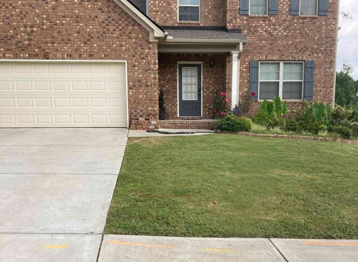 Front view of a brick house with garage, front door, driveway, green lawn, bushes, flowers, and windows.