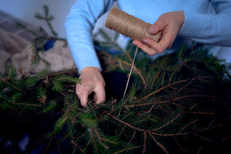 Hands work with pine branches while wrapping twine around clipped needles, preparing natural materials for crafting a handmade holiday wreath.