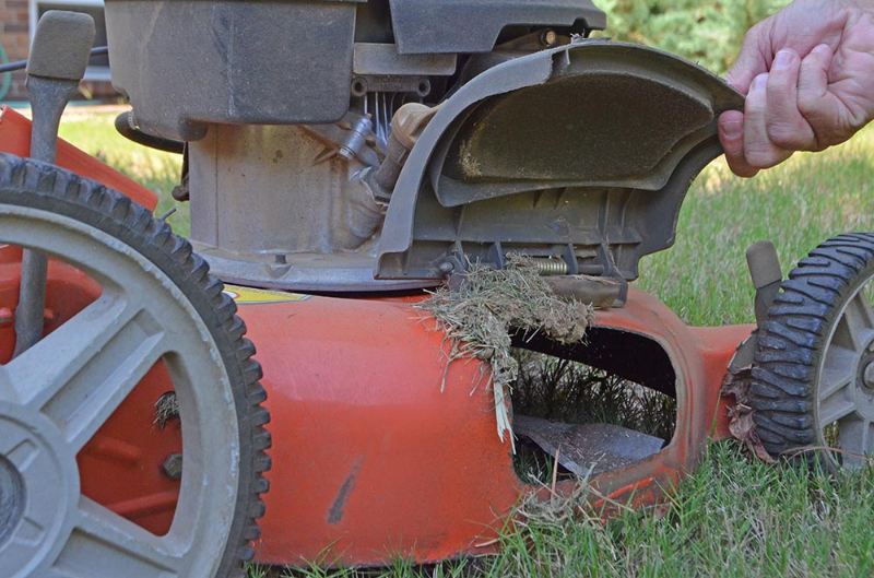Person lifting mower cover to reveal grass clippings stuck underneath deck, performing cleaning maintenance to prevent clogs and improve performance.