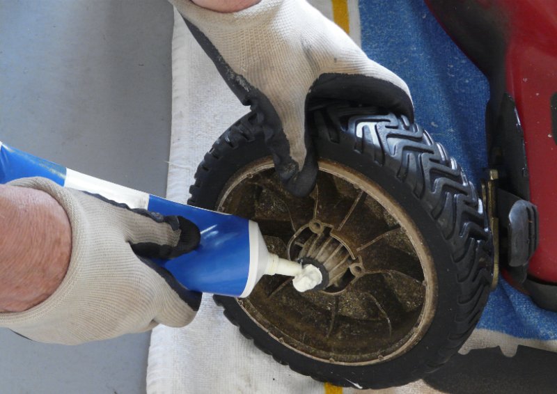 Person wearing gloves applying grease to lawn mower wheel bearing with a grease gun, performing maintenance to ensure smooth operation.