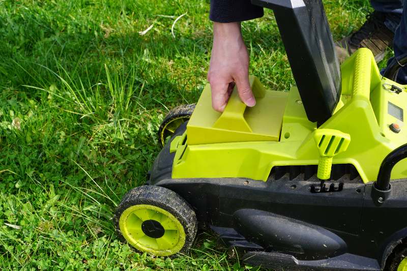 Person removing rechargeable battery pack from bright green electric lawn mower on grass, ensuring safety and proper storage after mowing is complete.