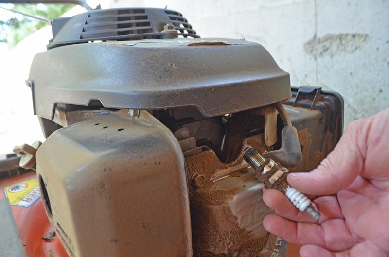 Hand holding dirty spark plug removed from lawn mower engine, showing essential maintenance step for reliable ignition and performance. 