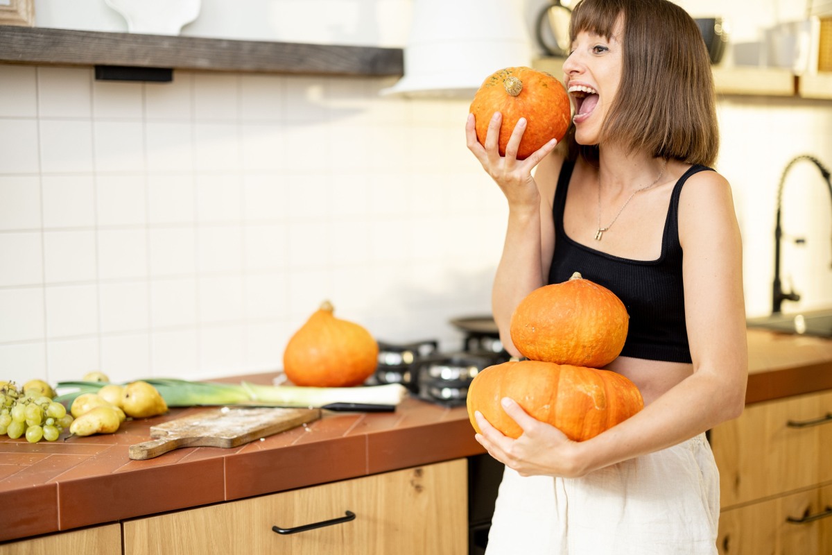 Woman in the kitchen holding pumpkins in her left hand and biting one with her right hand