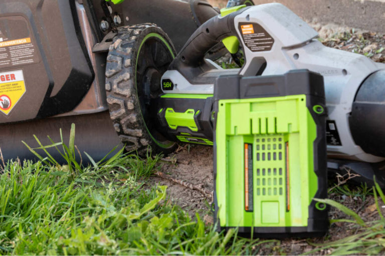 Green 56V battery pack sits beside cordless yard tool near lawn mower wheel on grass, ready for outdoor work.