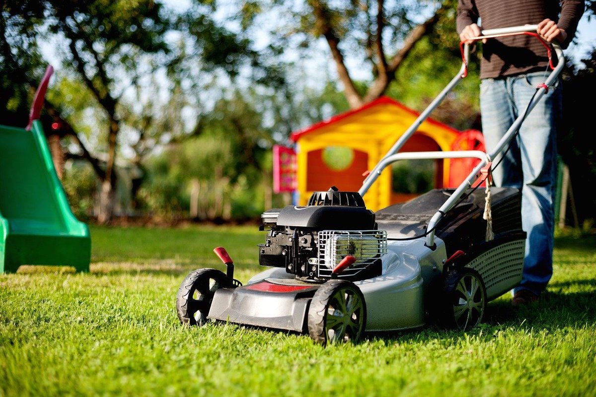 Man mowing a lawn mower with a swing set slide and a child's playhouse in the background.