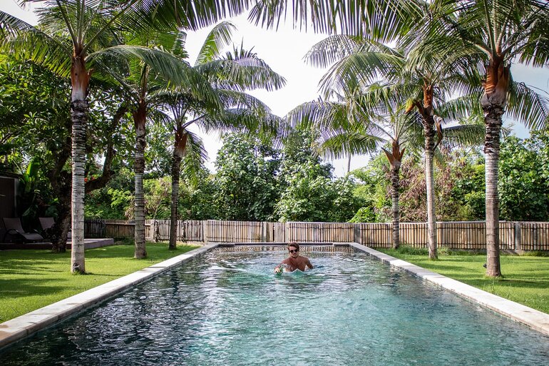 Man swimming in inground pool lined on both sides with palm trees