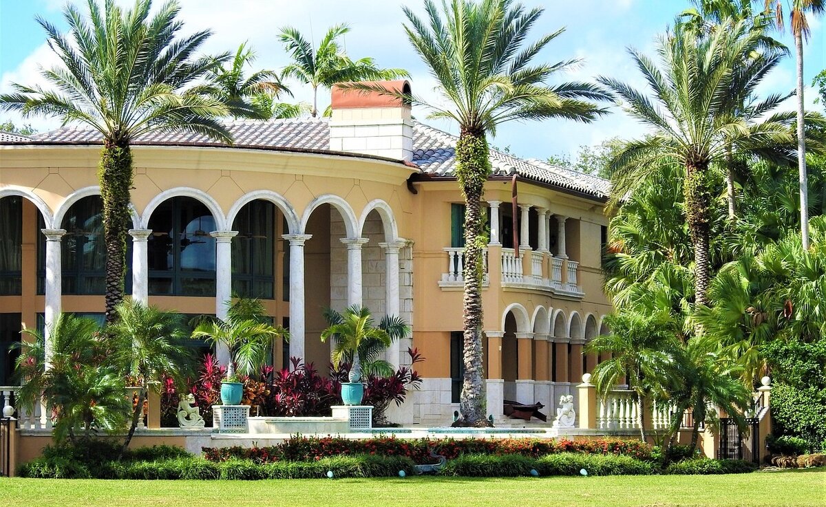 Large yellow house with white stone columns and palm trees of different sizes all around it