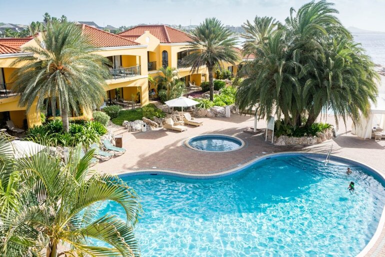 A pool and hot tub behind a resort with a clump of palm trees casting shade