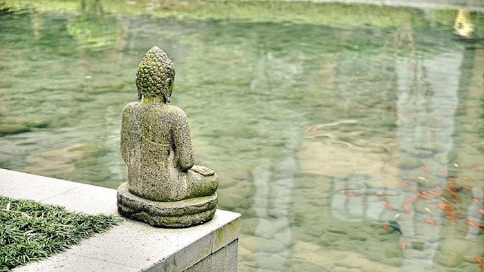 Buddha sculpture sitting on edge of platform, facing clear water