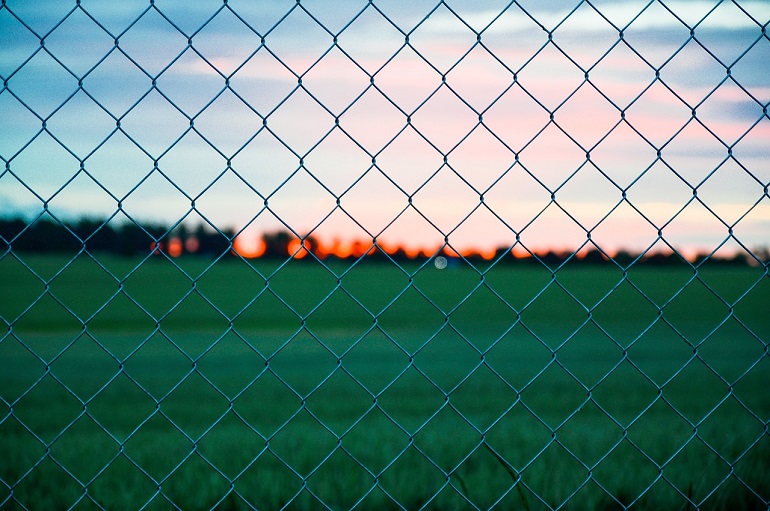 Close up of metal chain link fence in front of green field and sunset