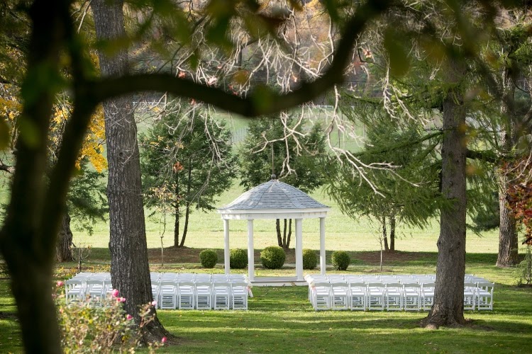 White, wedding gazebo in semi-wooded field with dozens of white guest chairs