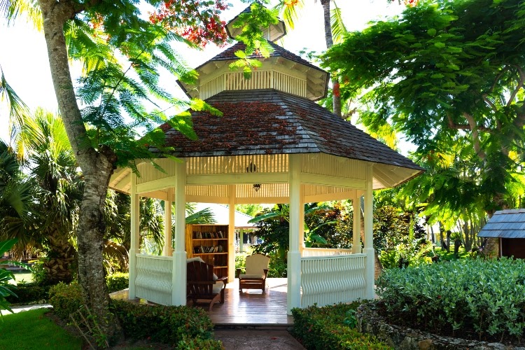 White gazebo with bookshelf and two lounge chairs, surrounded by low shrubs and tall trees