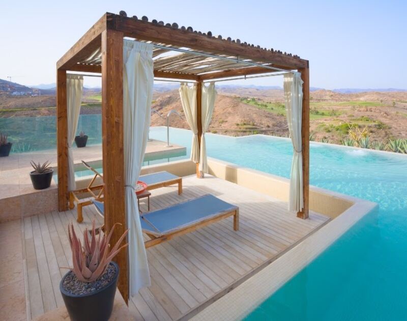 wooden, poolside pergola with white curtains on a wooden pool deck above lounge chairs