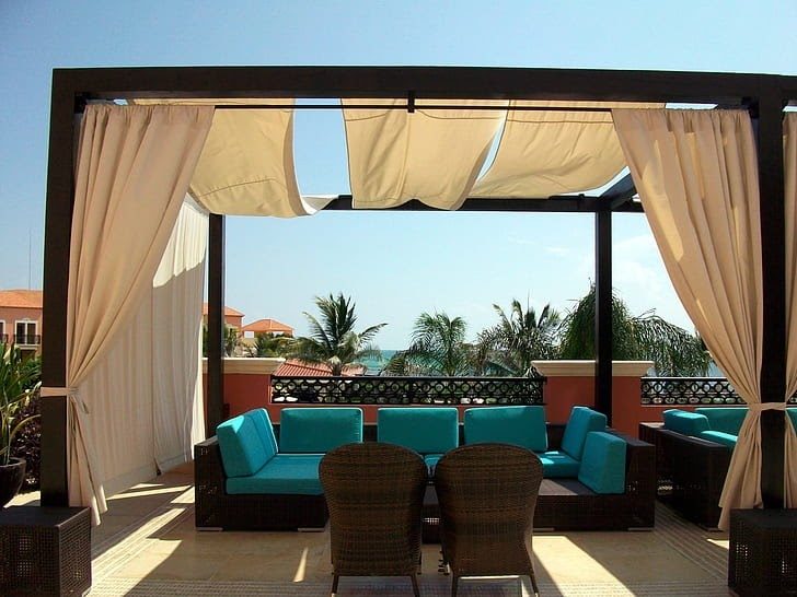 brown pergola with cream-colored curtains over patio with brown and teal chairs