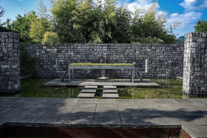 Six stepping stones lead to small seating area in the middle of a large green pond