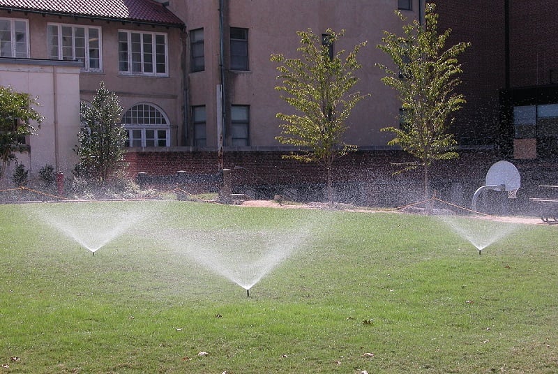 three sprinklers watering a lawn with building and trees in the background