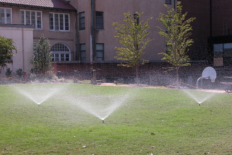 three sprinklers watering a lawn with building and trees in the background