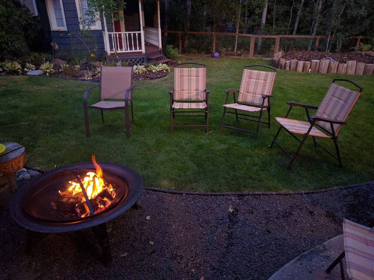 View of a backyard with a fire pit in the bottom left corner, 4 lawn chairs on the lawn, and a house in the background