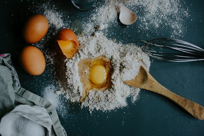 A flat lay of baking materials, including flour, raw eggs, a mixing spoon, and a whisk