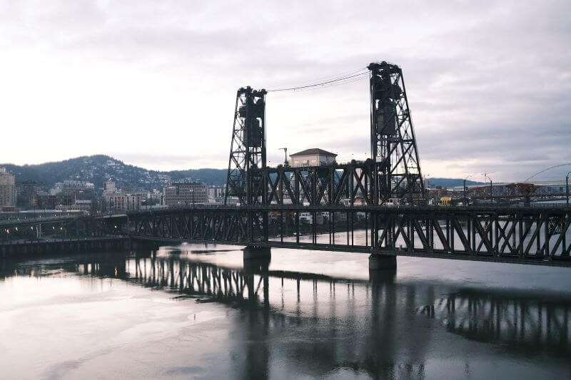 A drawbridge reflects on the surface of a river, in front of buildings emerging across Portland’s landscape on an overcast day.