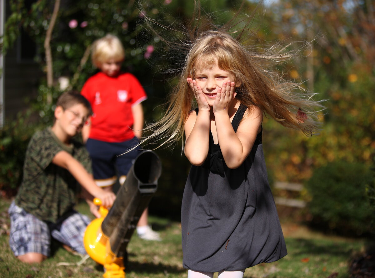Young boys aiming a leaf blower at a young girl, and her hair is blown upwards