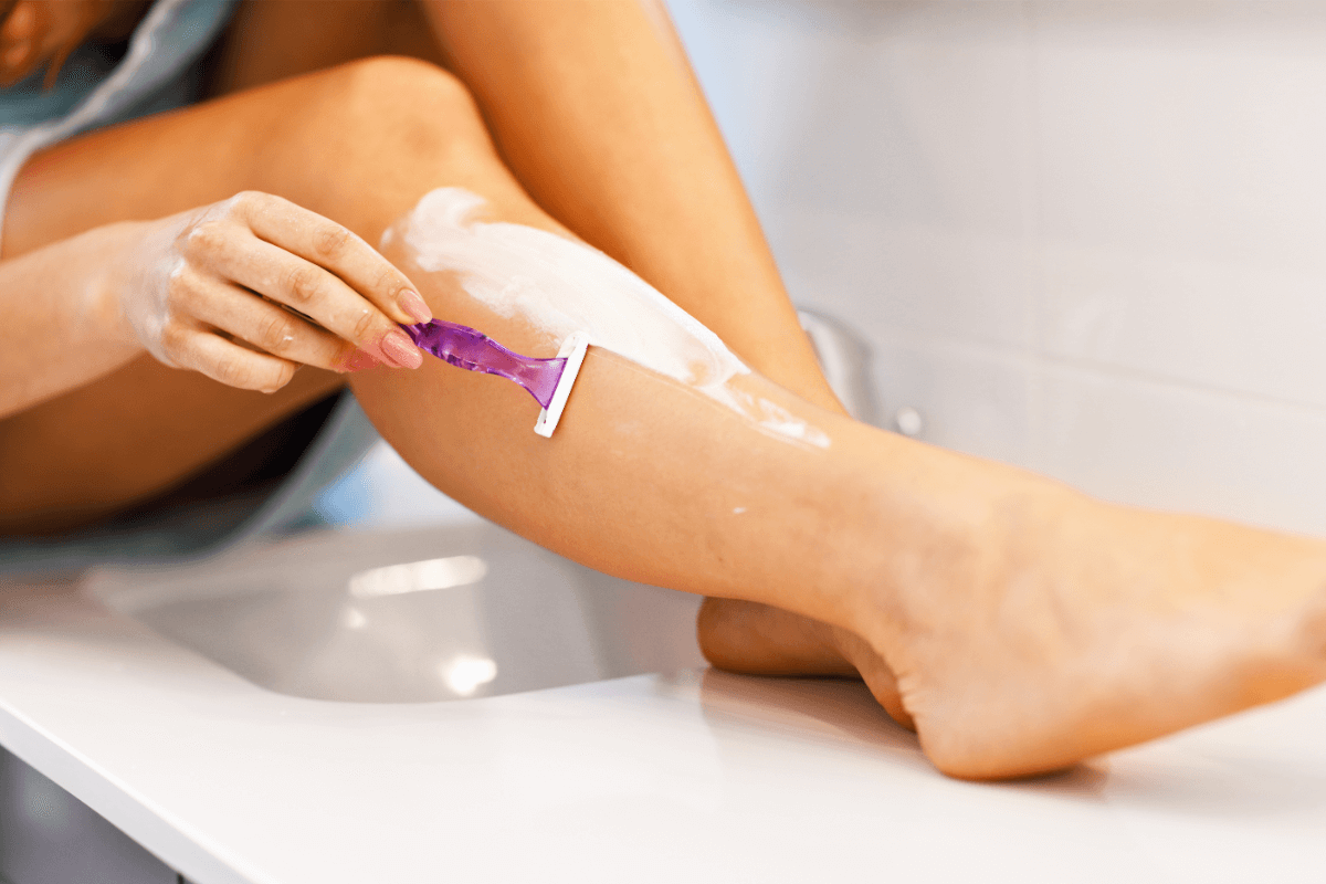 A woman uses a razor to shave her legs covered in shaving cream over the sink.
