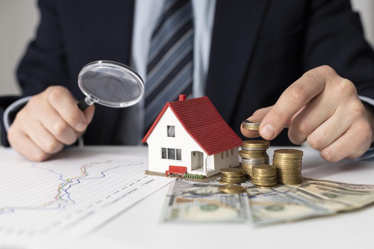 Photo illustration of a man with a tiny house model on his desk with paper and coin money beside it. He is holding a magnifying glass.