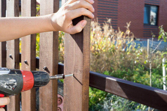 A fence being built with one hand bracing the fence as another hand wields a power screwdriver to secure a plank to the fence frame.