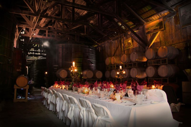 A long, candlelit table in the middle of the Bernardo Winery barrel room is decorated with a white tablecloth and surrounded by white-fabric-covered chairs and awaits guests