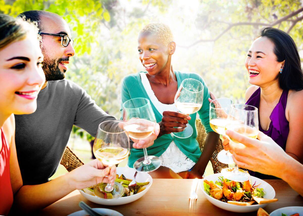 A group of friends sitting around a table in a vineyard toast their wine glasses