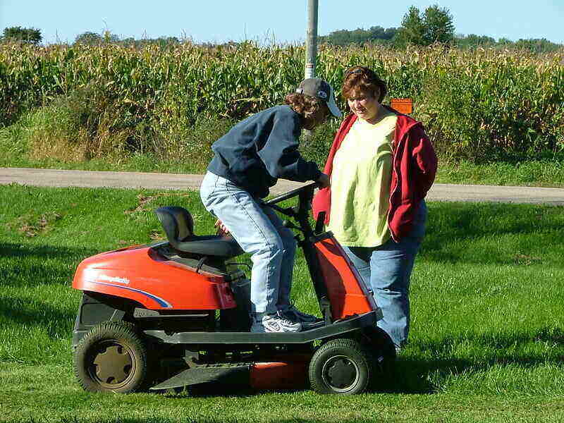 Rear-engine lawn mower with a woman sitting on it and another woman standing next to it