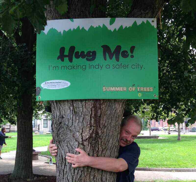 Man hugging a tree with a sign on the tree that says, "Hug Me! I'm making Indy a safer city."
