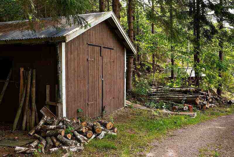 Brown, wood shed with a metal roof nestled into the woods with firewood on either side of it