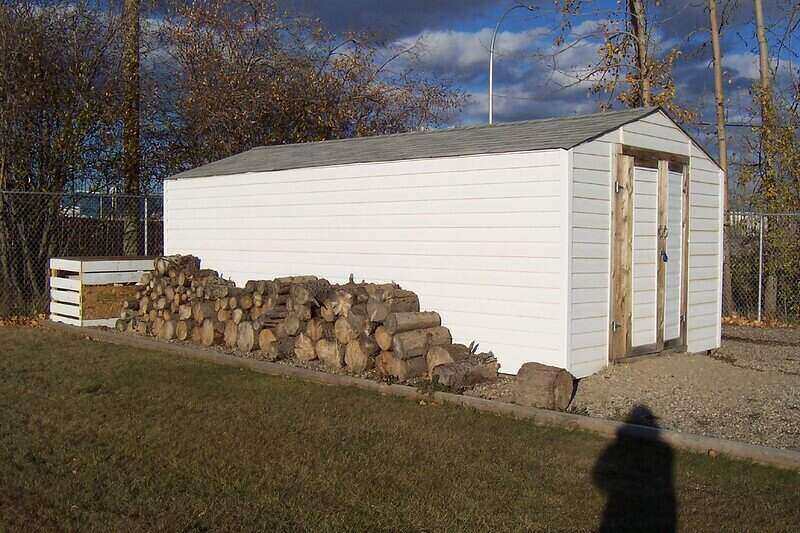 Large white shed with a pile of firewood stacked along the side of it