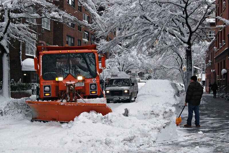 Snow plow on the street and a man holding a shovel on the sidewalk