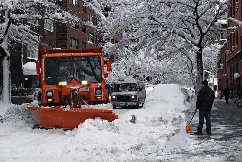Snow plow on the street and a man holding a shovel on the sidewalk