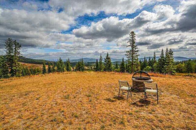 Open field, clouds, trees, mountains in distance with two chairs and a fire pit in the foreground