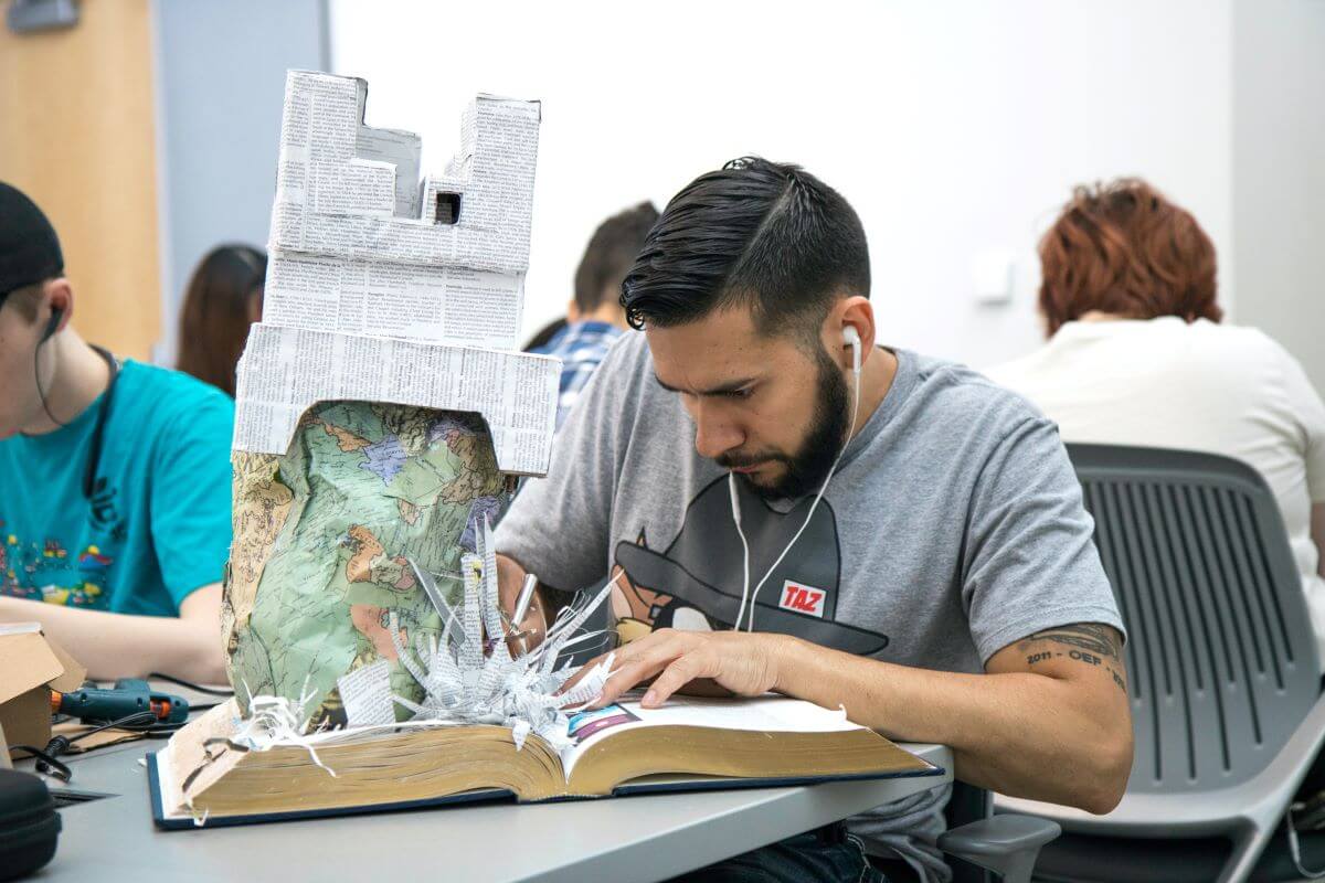 A man sits among a group of people as he tears out pages out of a big book to create a towering architectural sculpture.