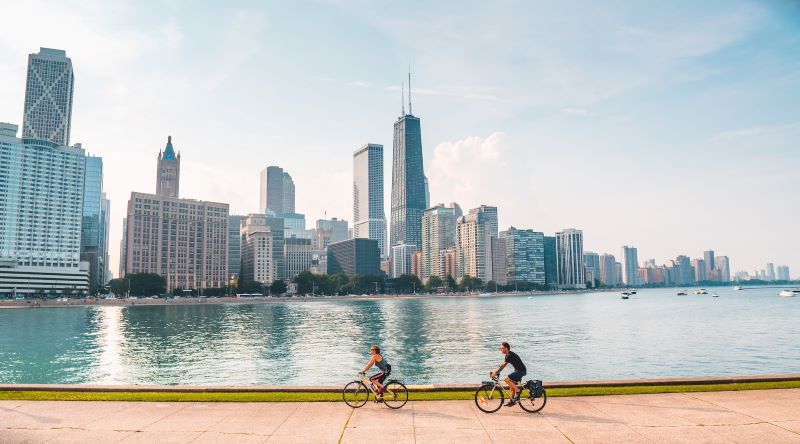 Two people bike along the river across from the Chicago skyline.