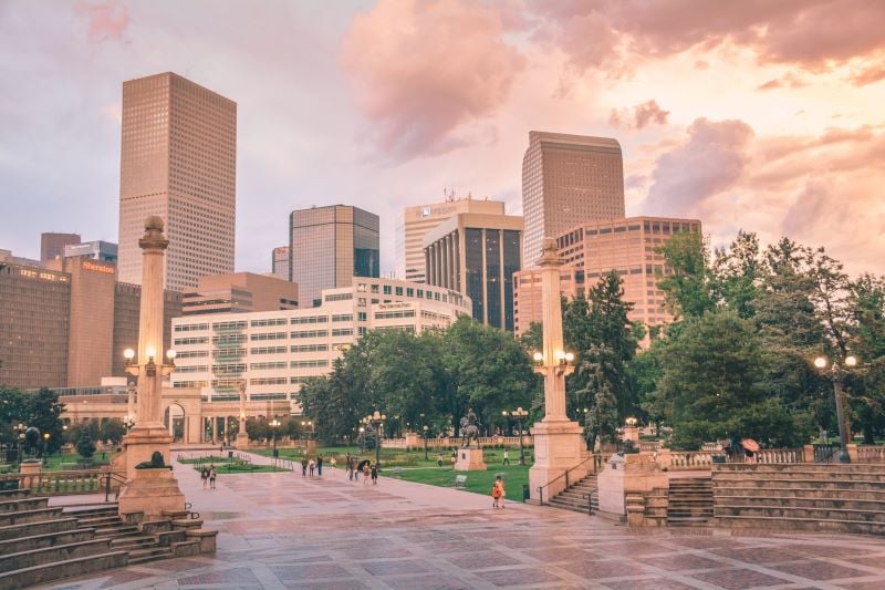 People walk around a park in Denver while skyscrapers in the background reflect the sunshine emerging from a break in the cloudy sky.