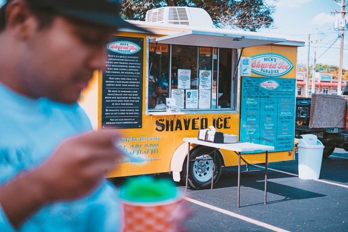 A man in the foreground enjoys his shaved ice. In the background is Nick’s Shaved Iced Paradise food truck in a parking lot.