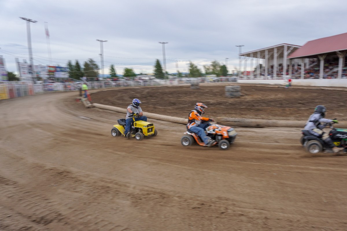 Three racers on riding lawn mowers racing around a dirt track at a fairground